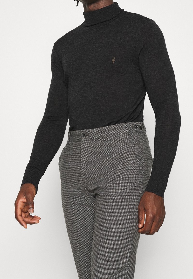 Schwarzer Rollkragenpullover aus glattem Material mit einem dezenten Logo. Kombiniert mit gemusterten grauen Hosen, die eine taillierte Passform haben.