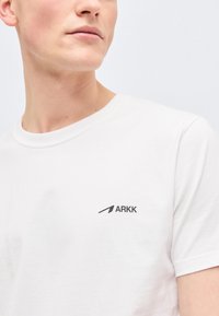 Νέος άνδρας που φοράει απλό λευκό t-shirt με μικρό μαύρο λογότυπο ARKK στο αριστερό στήθος, κοιτάζοντας προς τα πλάγια μπροστά σε απλό φόντο.