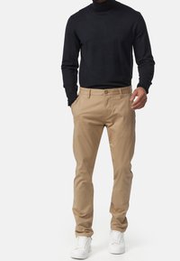 Man med beige chinos, svart långärmad polotröja och vita sneakers, stående med en hand i fickan.