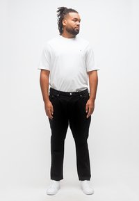 Άνδρας με κοτσίδες ντραντλόκ, φοράει λευκό T-shirt, μαύρο παντελόνι και λευκά αθλητικά παπούτσια, στέκεται μπροστά σε μια απλή λευκή επιφάνεια, κοιτάζοντας προς τα πλάγια.