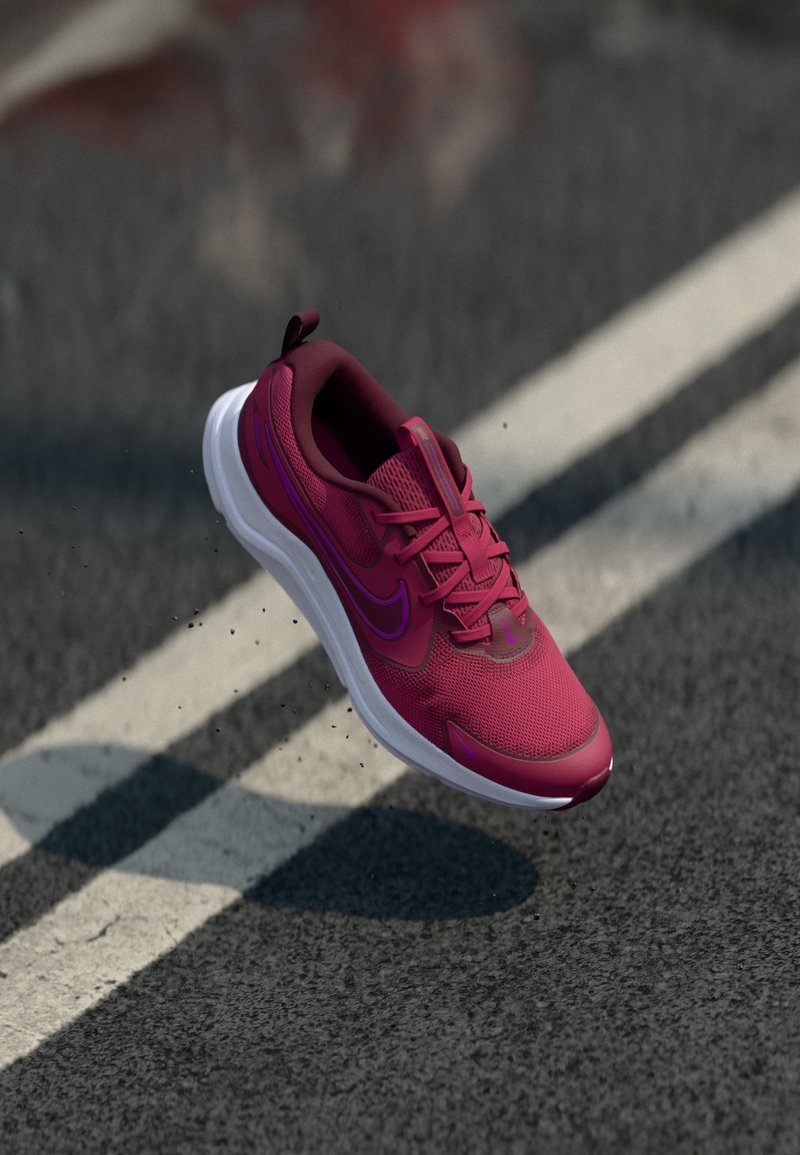 Shoe desportivo rosa com material em malha, detalhes contrastantes em roxo, design leve e sola de borracha branca, flutuando acima de uma superfície pavimentada.
