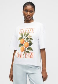 Γυναίκα με πλεγμένα μαλλιά που φοράει μια λευκή υπερμεγέθη μπλούζα με πορτοκάλι και το κείμενο «ELLESSE DAL 1959», συνδυασμένη με ανοιχτό μπλε παντελόνι.