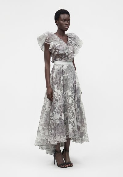 Γυναίκα φοράει ένα γκρι διάφανο φόρεμα με φλοράλ σχέδιο, βολάν στα κοντά μανίκια και φούστα με ασύμμετρο τελείωμα, συνδυασμένο με μαύρα ψηλοτάκουνα με λουράκι στον αστράγαλο.