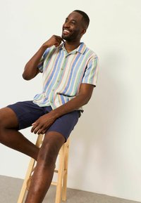 Homme souriant portant une chemise à rayures pastel à manches courtes et un short bleu marine, assis sur un tabouret en bois devant un fond uni.
