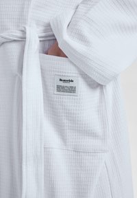Bata blanca de textura de waffle con cinturón en la cintura y bolsillo. Una etiqueta en el interior dice "Resteröds", indicando la marca o el origen.