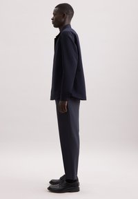 Granatowa koszula z długim rękawem i kołnierzykiem, zestawiona z dopasowanymi spodniami i czarnymi eleganckimi butami, widok z boku.