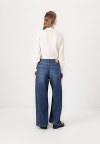Jeans typu wide-leg w ciemnoniebieskim denimu, z wysokim stanem, tylne kieszenie i beżową metką na pasie. Zestawione z jasną bluzką.