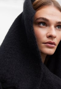 Svart ullkappa med en mjuk, ludden yta som hänger över huvudet och axlarna, och visar en minimalistisk design och djup färgton.