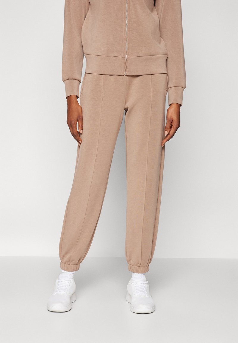Pantalons de jogging beige en tissu doux, dotés d'une taille élastique et de poignets, avec des coutures verticales pour plus de détails, associés à des baskets blanches.