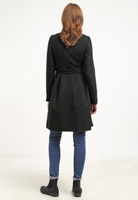 Schwarzer Mantel mit figurnaher Passform, tailliertem Gürtel, langen Ärmeln und Schlitzen im Rücken; kombiniert mit blauen Jeans und Stiefeletten.