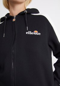 Personne portant un sweat à capuche noir zippé avec des bandes blanches sur les manches et le logo "ellesse" sur la poitrine ainsi que sur la tirette de la fermeture éclair.