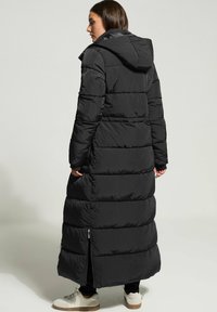 Svart pufferjacka med lång design, med dragkedjeaccent och huva. Tillverkad av quiltat, texturerat tyg med en matt finish.