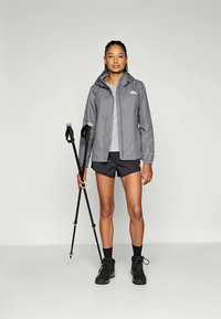 Lichte grijze jack met capuchon, voorklinken en logo; gecombineerd met een grijze top en zwarte shorts; houdt wandelstokken vast in een neutrale setting.