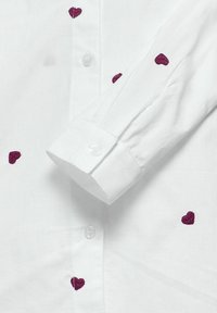 Camisa de algodón blanca con mangas largas, que presenta corazones bordados en púrpura y cierre de botones en la parte delantera. Textura suave y diseño de cuello clásico.