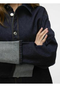 Denimjacka med mörkblått tyg och ljusgrå ärmar. Designen inkluderar krage, knappstängning och synliga sömsdetaljer.