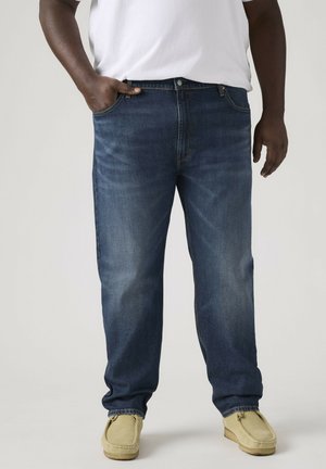 Volwassene die blauwe spijkerbroek, wit T-shirt en beige loafers draagt, staand met de linkerhand in de zak tegen een effen achtergrond.