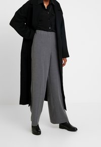 Persona vestida con abrigo negro, camiseta negra, pantalones anchos grises y botines negros, de pie contra un fondo liso de color gris claro.