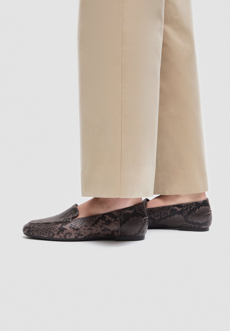 Personne portant un pantalon beige et des mocassins marron à motif peau de serpent, debout sur un fond uni clair.