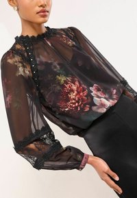 Priehľadná čierna blúzka s kvetinovými vzormi v červenej a zelenej farbe, čipkovými detailmi na golieri a rukávoch a priliehavým čiernym pásom.