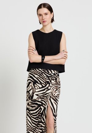 Femme aux cheveux courts et foncés portant un haut noir sans manches et une jupe imprimée zèbre avec une fente latérale, debout les bras croisés.