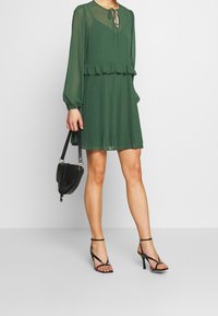 Vestido de gasa verde con mangas largas, cintura voluminosa y falda plisada. Accesorios incluyen un bolso negro y sandalias de tacón alto con tiras.