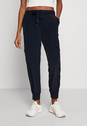 Person trägt marineblaue Cargo-Jogginghosen mit elastischen Bündchen und weiße Sneakers, steht vor einem schlichten Hintergrund.