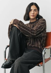 Brązowy sweter z białymi pionowymi paskami, w połączeniu z luźnymi, ciemnymi dżinsami. Model siedzi na chromowanym krześle.