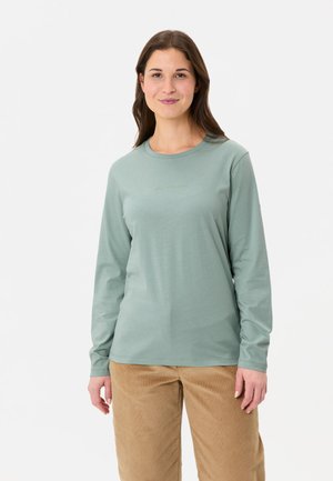 Langärmliges, hellgrünes Hemd aus weichem Material mit rundem Halsausschnitt und dezentem Branding auf der Brust, kombiniert mit beigefarbenen Cordhosen.