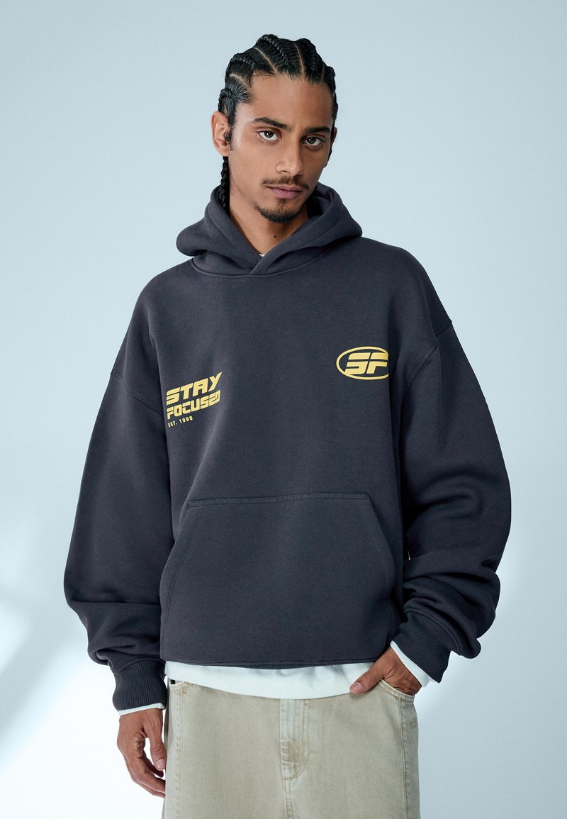 Homme aux cheveux tressés portant un sweat à capuche noir avec le texte jaune « Reste concentré » et un pantalon beige, debout devant un fond bleu clair.