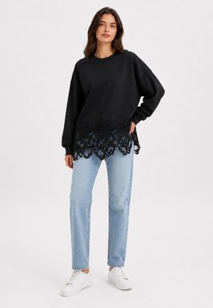 Jonge vrouw staand, draagt een zwart topje met lange mouwen en een kanten zoom, lichtblauwe spijkerbroek en witte sneakers op een effen witte achtergrond.