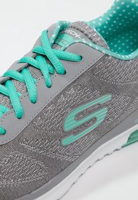 Baskets de sport grises avec un matériau en maille texturée, lacets turquoise et logo. Présente une semelle légère et une doublure intérieure à pois.