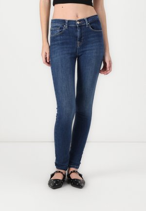 Kvinde iført mørkeblå skinny jeans og sorte flade sko med sølvfarvede nitter, stående mod en ensfarvet hvid baggrund.