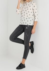 Leichte Cremebluse mit grauen Punkten, kombiniert mit schwarzen Skinny Jeans mit ausgefransten Säumen und distressed Details. Schwarze Schnürschuhe.