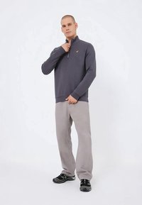 Vīrietis, kurš ģērbjas tumši pelēkā ceturtdaļas rāvējslēdzēja džemperī, gaiši pelēkās biksēs un melnās kedās, stāv uz viendabīga balta fona.