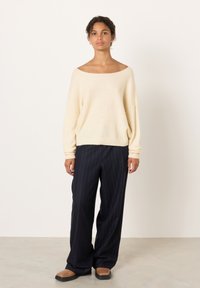 Sieviete ar cirtainiem matiem krēmkrāsas plecus atklājošā džemperī, tumši zilu plānu svītru biksēs ar plašām kājām un brūnās kurpēs ar kvadrātveida purngalu stāv uz vienkārša fona.