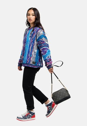 Woman wearing a colorful blue patterned sweater, black pants, red and blue high-top sneakers, holding a black patterned handbag by its strap.