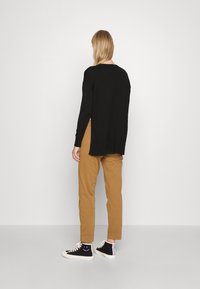 Personne aux longs cheveux blonds portant un pull noir, un pantalon marron et des baskets montantes noires, se tenant face à un mur blanc uni.