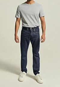 Homme portant un t-shirt gris clair à manches courtes, un jean bleu foncé et des baskets blanches, debout devant un fond uni.