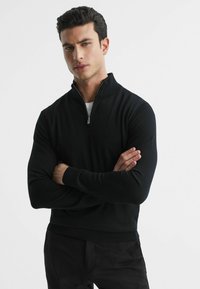 Pulover negru cu fermoar, cu mâneci lungi, tivuri și manșete ribbate, realizat dintr-un material moale și texturat. Modelul stă cu brațele încrucișate.