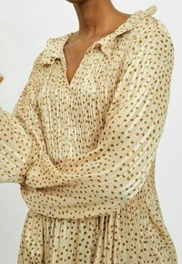 Blusa beige con colletto, caratterizzata da righe verticali e motivi a pois dorati. Tessuto fluido con superficie testurizzata e maniche a sbuffo.