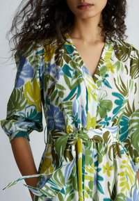 Vestido con estampado floral que presenta hojas verdes, azules y amarillas. Diseño envolvente con cintura ajustada, escote en V y detalles de botones. Tela suave.