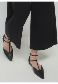 Piernas vestidas con pantalones de pierna ancha negros y zapatos planos negros de punta con hebillas en la tobillo tipo T sobre un suelo claro.