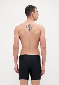 Čierne plavecké šortky vyrobené z hladkého, elastického materiálu, s elegantným dizajnom a viditeľným pásom, nosené modelom zozadu.