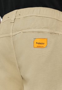 Beige pants with an elastic waistband and a back pocket featuring an orange Replay Inc. label worn with a black top.