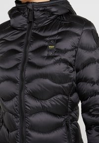 Schwarze Pufferjacke mit Kapuze, versehen mit Wellenmuster-Steppung, einem Frontreißverschluss und einem Logopatch auf der linken Brustseite.
