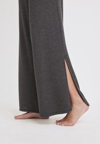 Graue, gerippte, weit geschnittene Hosen mit seitlichen Schlitzen. Weiche Textur, verlängerte Form und saubere Säume. Barfuß neben den Hosen sichtbar.