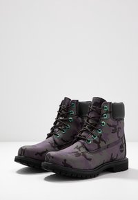 Botas de camuflaje moradas con acentos negros, parte superior de cuero texturizado, ojales turquesa en contraste y suela de goma negra resistente.
