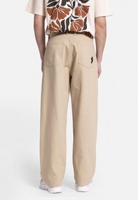 Pantalones de pierna ancha beige con bolsillos traseros, con un pequeño bordado de cactus. Llevados con una camisa de estampado floral, modelo de espaldas.