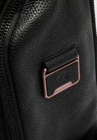 Bolso de cuero negro texturizado que presenta una cremallera prominente y una placa metálica con un logotipo en relieve, mostrando un diseño elegante y moderno.