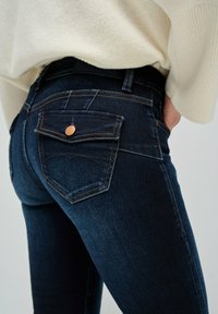 Vaqueros de mezclilla azul oscuro con un diseño ajustado, que cuentan con herrajes de cobre, costuras en contraste y un detalle de doble bolsillo en la parte posterior.